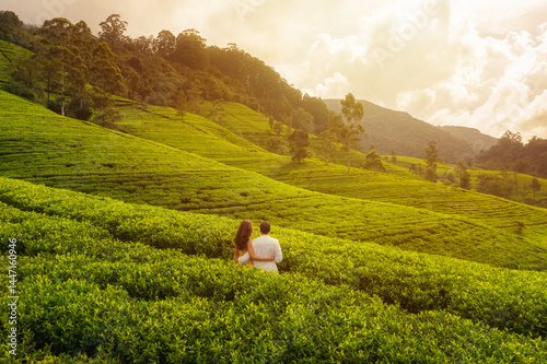 Obraz na płótnie A couple stands with their backs to the camera, admiring the vibrant tea fields of Sri Lanka during a golden sunset. This serene scene is perfect for nature, travel, and romance-themed visuals. w salonie