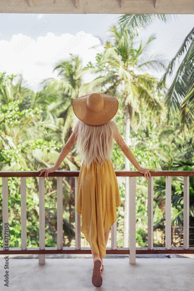 A stylish woman in a yellow dress and sun hat stands on a balcony, gazing at the lush tropical landscape. Perfect for travel, vacation, and lifestyle themes, evoking relaxation and wanderlust.