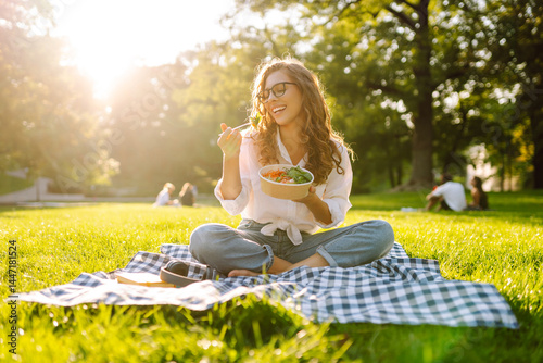Foto Happy woman in glasses sits on a mat in a park and eats a green salad