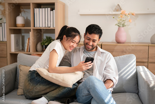 Portrait young Asian couple enjoy watching content on mobile phone together on cozy sofa, laughing with happiness in living room at home
