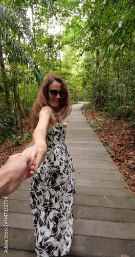 Happy tourist woman wearing dress holds her partner's hand and guides playfully through lush green park in Singapore. Wooden path in tropical nature with scenic atmosphere