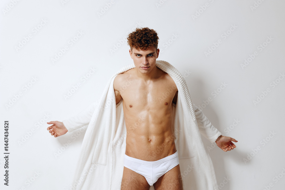 Fototapeta premium Confident young man in white underwear posing with an open white robe, showcasing his physique against a light background. Fashion and body positivity concept.