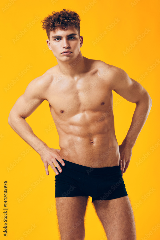 Fototapeta premium Athletic young man posing confidently in black swim shorts, showcasing toned physique against a bright yellow background. Fitness and health concept.