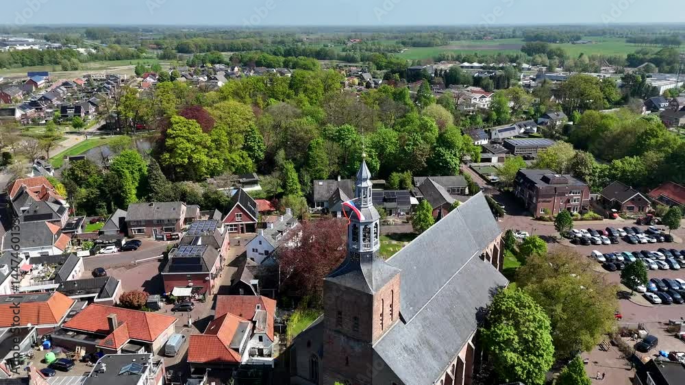 Historic church of Groenlo with waving durch flag on sunny day. People on streets celebrating Kings Day in Netherlands. Spring day with green and red colored trees. Aerial orbit shot.