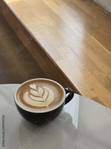beautifully crafted latte with intricate foam art sits in black cup on sleek surface, surrounded by warm wooden tones, creating cozy atmosphere