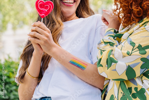 Fotografie Lesbian couple embracing, showcasing rainbow tattoo and heart lollipops, express