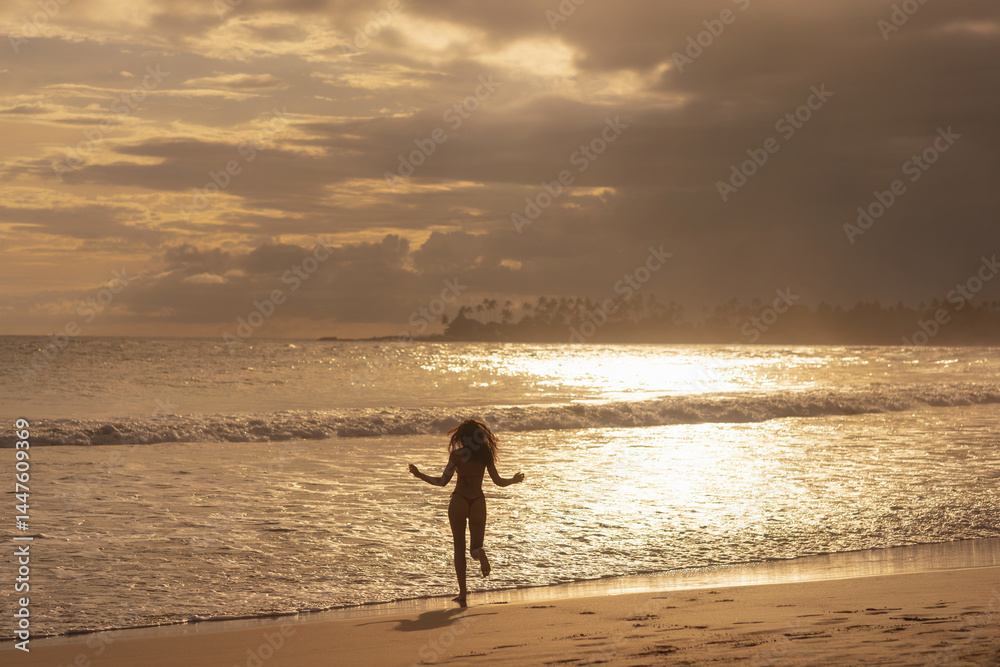 Silhouette of a carefree woman running toward ocean waves during sunset on a tropical beach. A dreamy, peaceful moment ideal for travel, freedom, summer lifestyle and vacation-themed ads.