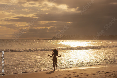Obraz na płótnie Silhouette of a carefree woman running toward ocean waves during sunset on a tropical beach. A dreamy, peaceful moment ideal for travel, freedom, summer lifestyle and vacation-themed ads. w salonie