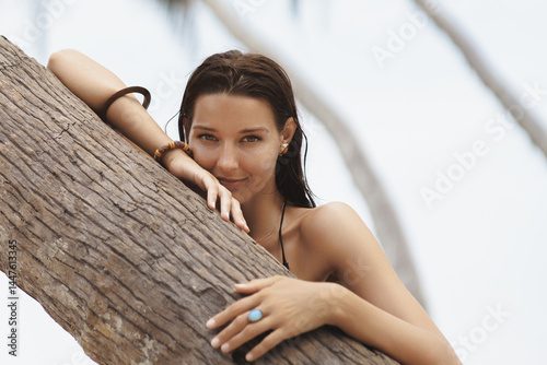 Obraz na płótnie Close-up of a confident Asian woman with stylish jewelry leaning on a palm tree by the beach. Ideal for beauty, wellness, spa, skincare, fashion, and tropical lifestyle advertising. w salonie