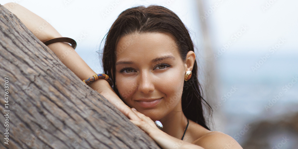 Close-up portrait of a tanned Asian woman resting on a palm tree with a gentle smile near the ocean. Perfect for beauty, skincare, spa, fashion, wellness and tropical lifestyle themes.