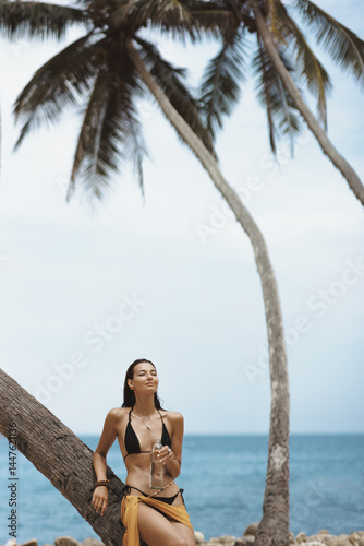 Obraz na płótnie Tanned Asian woman in black bikini with yellow sarong standing by palm tree, eyes closed, holding water bottle. Great for skincare, hydration, wellness, travel and tropical lifestyle promos. w salonie