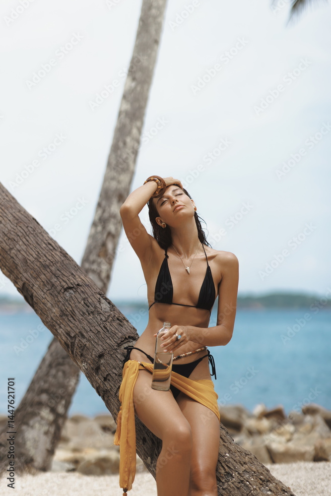 Smiling Asian woman in a black bikini and sarong holding a glass water bottle, enjoying sunshine by a palm tree on a tropical beach. Ideal for skincare, wellness and travel promo use.
