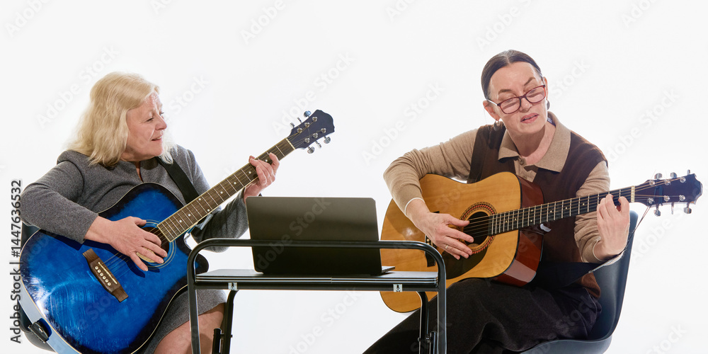 Obraz premium Elderly women playing acoustic guitars while studying music together with laptop during online lesson against white studio background. Concept of creative learning, education, friendship, aging.