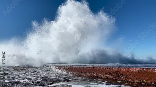 A giant wave hits an ocean rock wall with extreme force before exploding into a blue sky towards the camera. Captured in slow motion from in front of the the water explosion.
