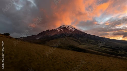 Snowcapped mountain peak rises amidst dramatic sunset clouds, creating a scenic landscape with brown grassy foreground hills.