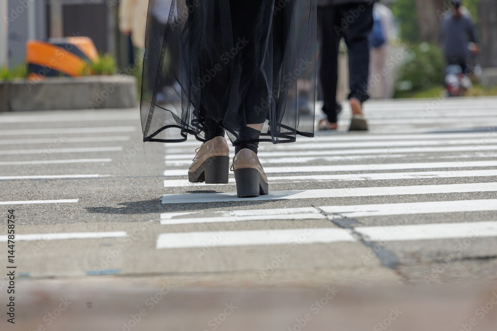 Low-angle person wearing light beige shoes and dark, sheer, black skirt-like garment walking across crosswalk marked by white painted lines on gray concrete. Angled perspective focuses on movement. Low-angle person wearing light beige shoes and dark, sheer, black skirt-like garment walking across crosswalk marked by white painted lines on gray concrete. Angled perspective focuses on movement.