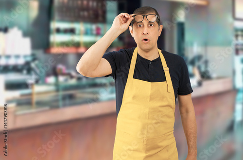 Mid adult with yellow apron straining his eyes because he can't see, at coffee shop photo