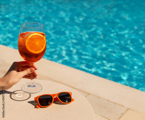 Aperol Spritz cocktail in a wine glass next to orange sunglasses on a poolside table, clear water and sunny atmosphere in background