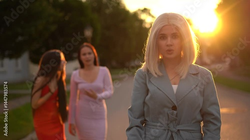 Wonderful Blonde girl stay and look at the camera, two girls talk in the background, sunset time
