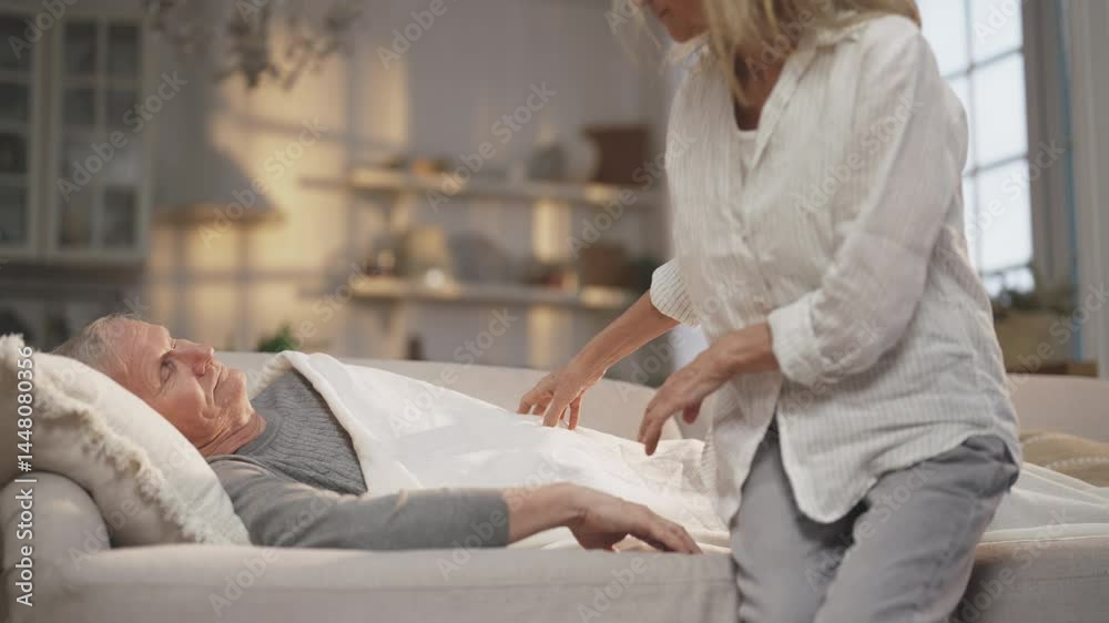 Sick elderly man lying on couch and careful wife wrapping him into blanket. Age spouses at home, husband feeling bad, catch cold in winter season, heart attack or hypertension, illness in old age