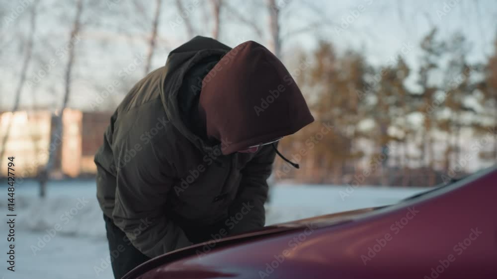 Man in hoodie bends over red car, struggling to open bonnet on snowy day with blurred view of residential buildings and bare trees in winter background