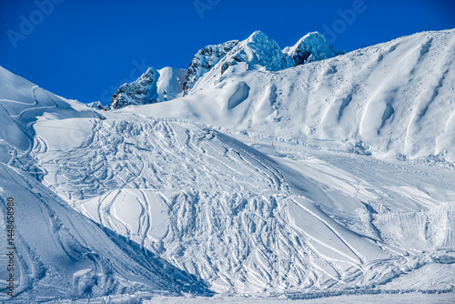 Fresh ski tracks create wavy patterns on a snow-covered slope in bright sunlight at a mountain resort during winter
