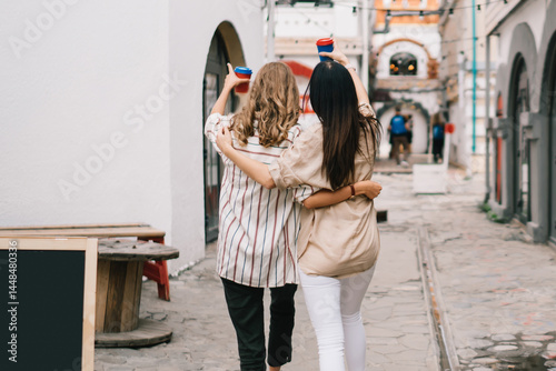 Back view of trendy dressed female friends with coffee to go exploring city urban setting and hugging,carefree women in casual clothing socialising during sincerely meeting in town for travel walking