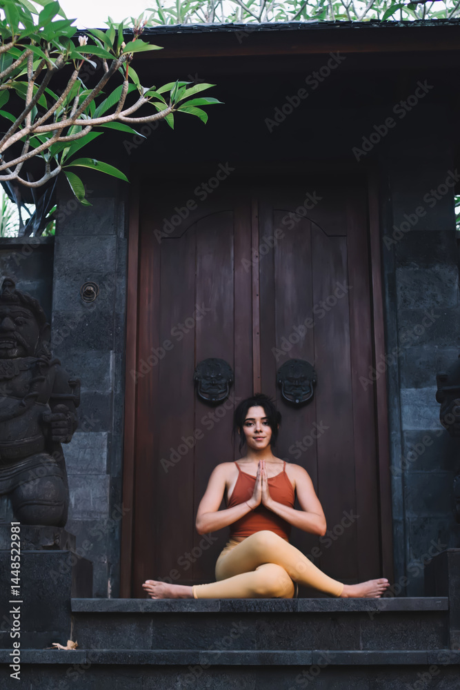 Young caucasian female in active wear sitting in yoga pose near door on temple satisfied with breathing and recreation, attractive woman looking in camera sitting in namaste asana enjoying harmony