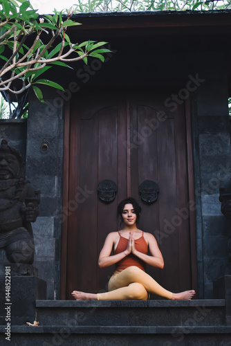 Obraz na płótnie Young caucasian female in active wear sitting in yoga pose near door on temple satisfied with breathing and recreation, attractive woman looking in camera sitting in namaste asana enjoying harmony w salonie