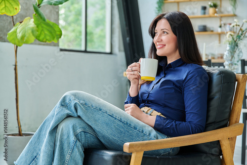 Portrait of beautiful 30s smiling woman with cup sitting in armchair at home in living room