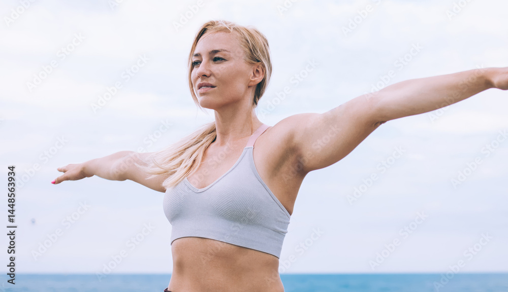 Caucasian female yogi practicing Virabhadrasana during morning workout at fresh ocean air, experienced slim woman in tracksuit doing warrior pose two enjoying healthy lifestyle for body harmony