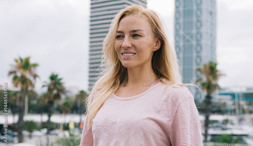 Carefree Caucasian female tourist dressed in casual clothing standing at urban setting and smiling, charming hipster girl in stylish outfit enjoying leisure time for visiting city during solo travel