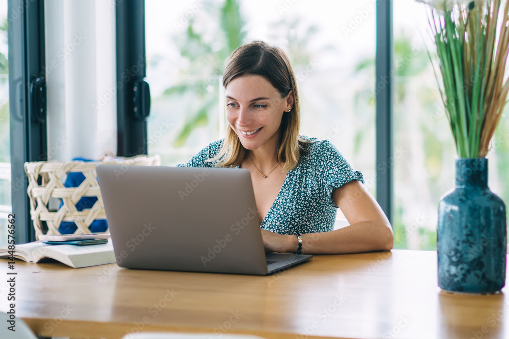 Smiling caucasian female digital nomad sitting at table in apartment satisfied with freelance job, happy clever 20s hipster girl learning online course and watching training webinar on laptop computer