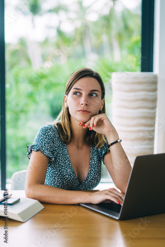 Thoughtful young woman working on laptop with window behind at home