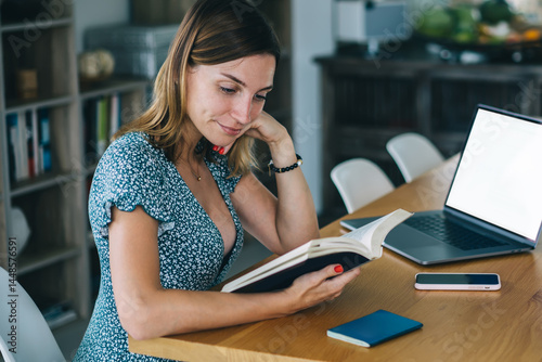 Positive millennial hipster girl enjoying interesting literature on free time sitting at apartment near laptop computer with mock up screen, smiling clever female student doing homework read textbook