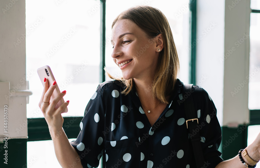 Positive millennial woman watching funny video on mobile phone enjoying coffee break in cafeteria, smiling carefree hipster girl millennial checking messages and mail via application on smartphone