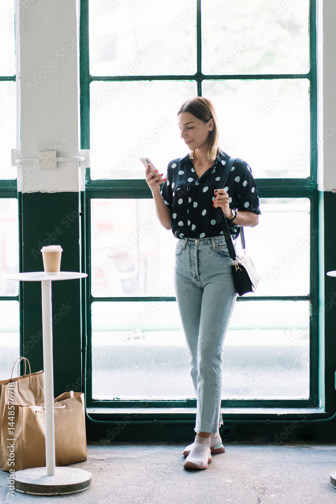 Young female in trendy wear using delivery service application on mobile phone getting shopping bags with purchases, young millennial woman reading income notification and messages on cellular