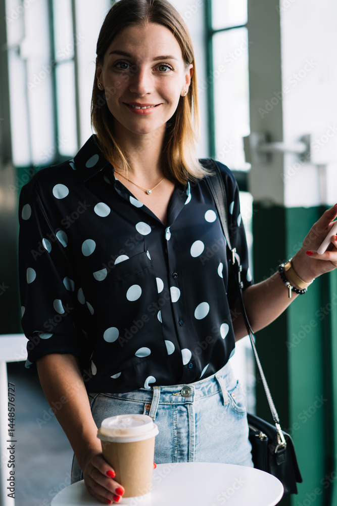 Half length portrait of smiling blonde female in trendy wear holding mobile phone and coffee cup on break, beautiful woman in blogger using smartphone for share media and online chatting in cafeteria