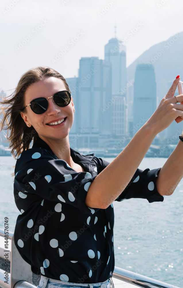 Happy caucasian female influencer shooting video for blog using mobile phone camera exploring downtown, smiling woman in sunglasses taking photos during vacation journey hold smartphone on bridge
