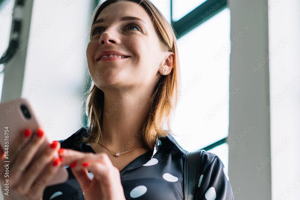 Smiling blonde female blogger influencer in trendy wear checking information on mobile phone indoors, positive woman 20s millennial holding smartphone making money transaction via app in cafeteria