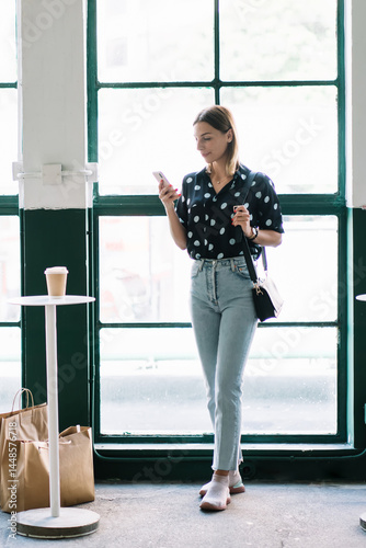 Obraz na płótnie Young female in trendy wear using delivery service application on mobile phone getting shopping bags with purchases, young millennial woman reading income notification and messages on cellular w salonie