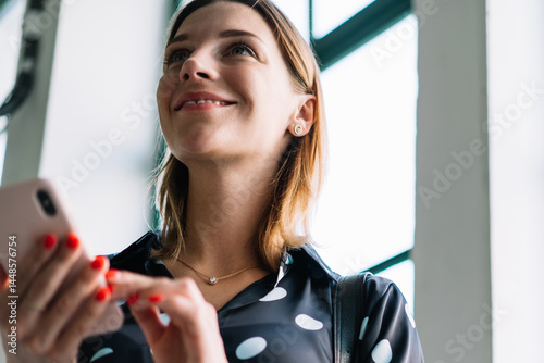 Obraz na płótnie Smiling blonde female blogger influencer in trendy wear checking information on mobile phone indoors, positive woman 20s millennial holding smartphone making money transaction via app in cafeteria w salonie