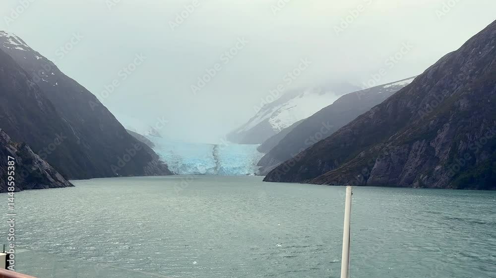 Misty view of a glacier flowing into a narrow fjord flanked by steep mountains, captured from a cruise ship under cloudy skies in southern Patagonia.