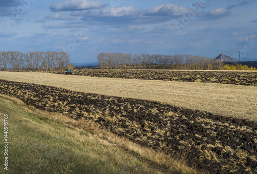 A tractor plows the land against the backdrop of a mountain and an industrial facility.