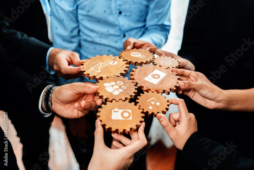 A group of diverse individuals collaboratively holds wooden gears featuring media icons, symbolizing teamwork and creative problem-solving in a professional environment. Amity