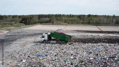 Dump truck unloading soil on a landfill site covered with mixed waste, surrounded by forest and infrastructure in the background.
 photo