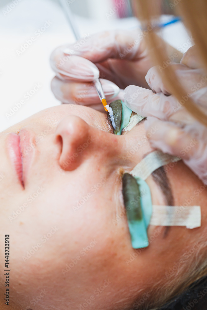 Eyelash lamination procedure on a woman's face. A cosmetologist glues long eyelashes with tweezers onto a woman's eye during an eyelash extension procedure.