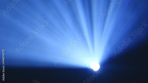 Bright blue spotlight projecting rays through haze or smoke, creating dramatic beams of light spreading outward in a dark environment with copy space.
