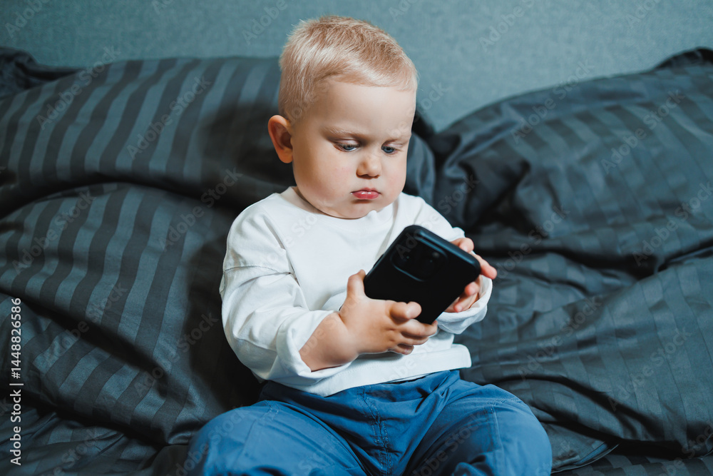Cheerful toddler boy sitting on bed and playing on smartphone during day at home. Close up of child sitting on bed holding smartphone at home. Smiling child with mobile.