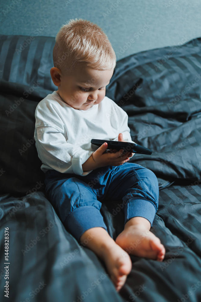 Cute little boy 2 years old using smartphone, looking at screen, child holding phone in hands, sitting on sofa at home alone, playing game on mobile device, watching cartoons online
